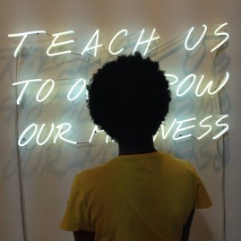 [Woman with Afro standing in front / obscuring sign that reads] Teach Us To Outgrow our Madness Alfredo Jaar